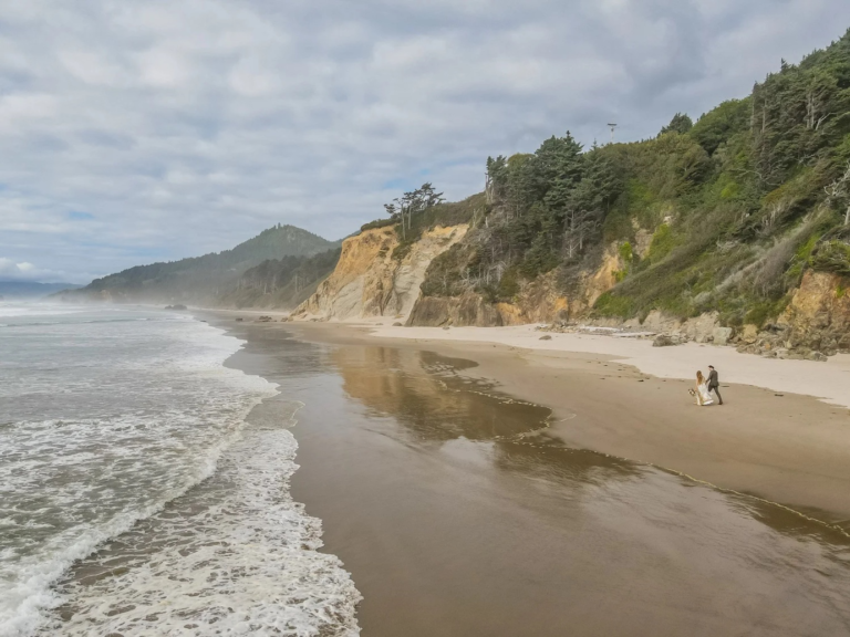Couple walking hand in hand along Arcadia Beach with sea stacks, sandy shoreline, and the Pacific Ocean in the background.